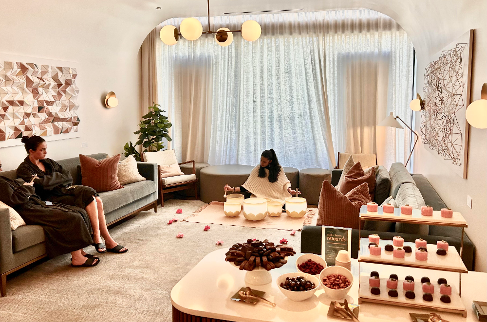 A cozy, upscale lounge or gift lounge setting with two women sitting on sofas, a coffee table with bowls of dried fruits and candles, and a display shelf of small pink and red packaged items. The room features warm neutral tones, beige curtains, and modern wall art.