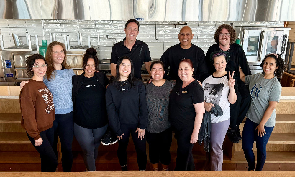 A group of ten people posing together in a modern kitchen or breakroom environment, wearing mostly black t-shirts.