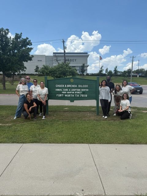 A group of ten people posing together outdoors in front of a green sign that reads 'Chuck & Brenda Gilcox Animal Care & Shifting Center Fort Worth TX 76109' under a blue sky with white clouds.