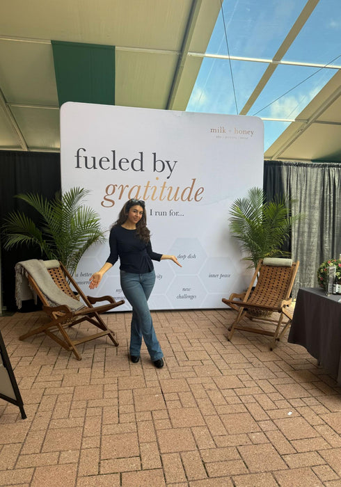A woman stands in front of a large white backdrop banner that reads 'fueled by gratitude' in black and gold lettering. She is wearing a dark navy long-sleeve top and blue jeans.