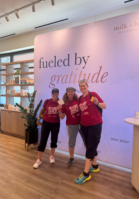 Three women standing together in front of a large pink and white 'milk + honey' branded wall inside a retail space, wearing matching red graphic t-shirts and posing with a small card.