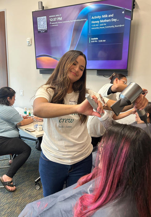 A woman in a salon setting uses a hair dryer on another person's pink-tinted hair, with other salon staff and a large event screen visible in the background.
