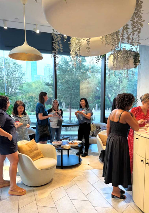 A group of women are gathered in a modern, stylishly decorated cafe or lounge space with large windows, pendant lights, and cream-colored furniture.