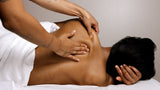 A person receiving a massage lies face down on a white treatment table while another person's tattooed arms apply pressure with their hands. The scene is shot from a high angle against a neutral gray background.