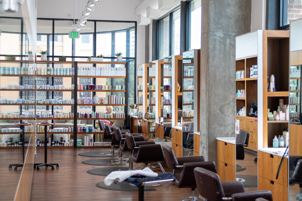 A modern hair salon interior with multiple salon chairs, wooden shelving filled with hair products, mirrors, and large windows.
