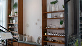 A styled interior scene showing wooden shelving units filled with small bottles, vases, and plants, alongside a cushioned bench with pillows in a bright room.