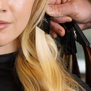 A close-up of a person with long blonde hair having a hair extension applied. A stylist's hand is holding a black fan of hair color samples next to the section of blonde hair being styled.