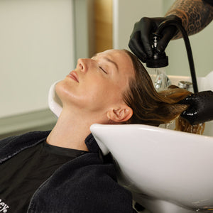 A woman with closed eyes reclines in a white salon shampoo bowl while another person wearing black gloves washes her hair. She wears a black salon cape with white text, and the setting appears to be a salon interior.