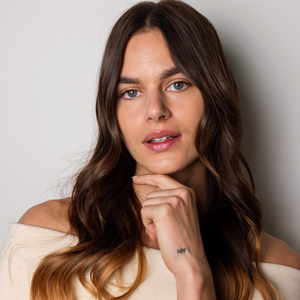 A woman with long, wavy brown hair with warm caramel highlights, wearing a cream off-the-shoulder top, posing against a neutral light gray background.