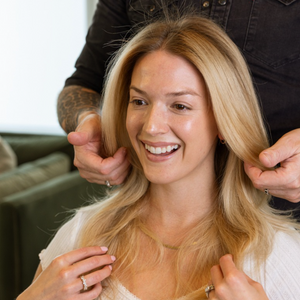 A smiling blonde woman sits while a tattooed stylist styles her hair. She wears a white top, and the background shows a softly blurred indoor setting with a green couch.