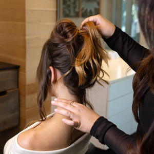 A woman with a messy bun hairstyle is shown from behind in a salon setting, while another person styles her hair. The hairstyle features dark brown hair with blonde highlights.