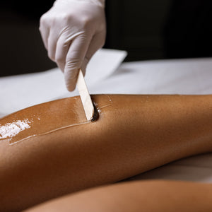 A close-up of a leg being treated with an adhesive hair removal gel, with a gloved hand using a spatula to spread it. The person's leg is resting on a white treatment table.