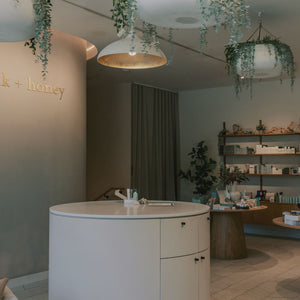A modern skincare or beauty retail interior with a round white counter, wooden display tables, shelves of products, hanging greenery, and warm pendant lights. The wall displays the brand name 'milk + honey' in gold lettering.
